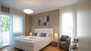 A modern bedroom with a large bed, two bedside tables with lamps, a chair, and a view of greenery outside.