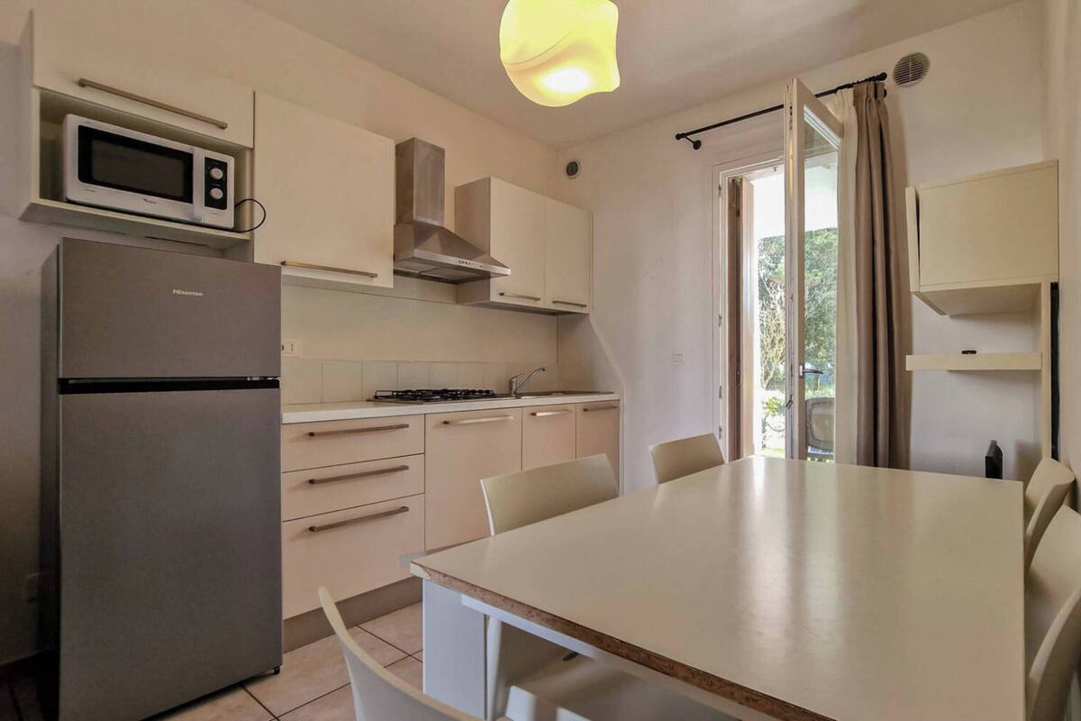 A compact kitchen with a refrigerator, microwave, stove, and white cabinets. There is a dining table with chairs and a window with curtains.
