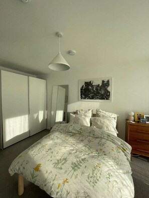 A bedroom with a bed, a bedside table, a wardrobe, a hanging light fixture, and a framed artwork on the wall.
