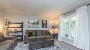 A living room with a sofa, a coffee table, a bookshelf, and a large window with curtains. - Woodford Stables / Walk to Eckert's Orchard (Versailles)