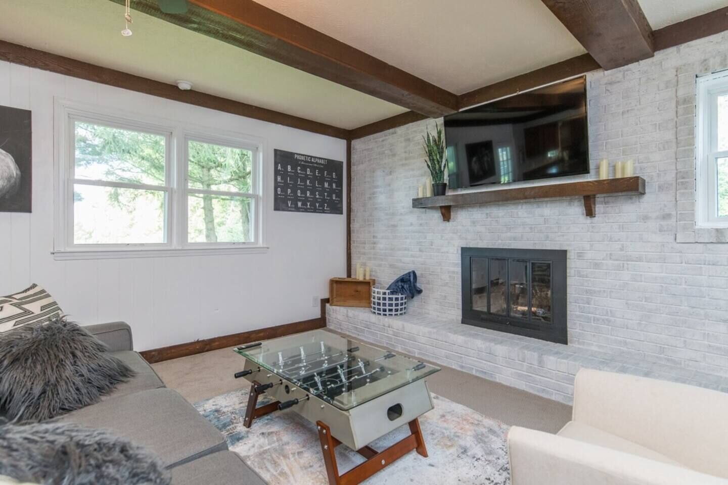 Una sala de estar con mesa de futbolín de vidrio, chimenea, televisor montado y un ventanal grande.