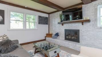 Una sala de estar con mesa de futbolín de vidrio, chimenea, televisor montado y un ventanal grande.