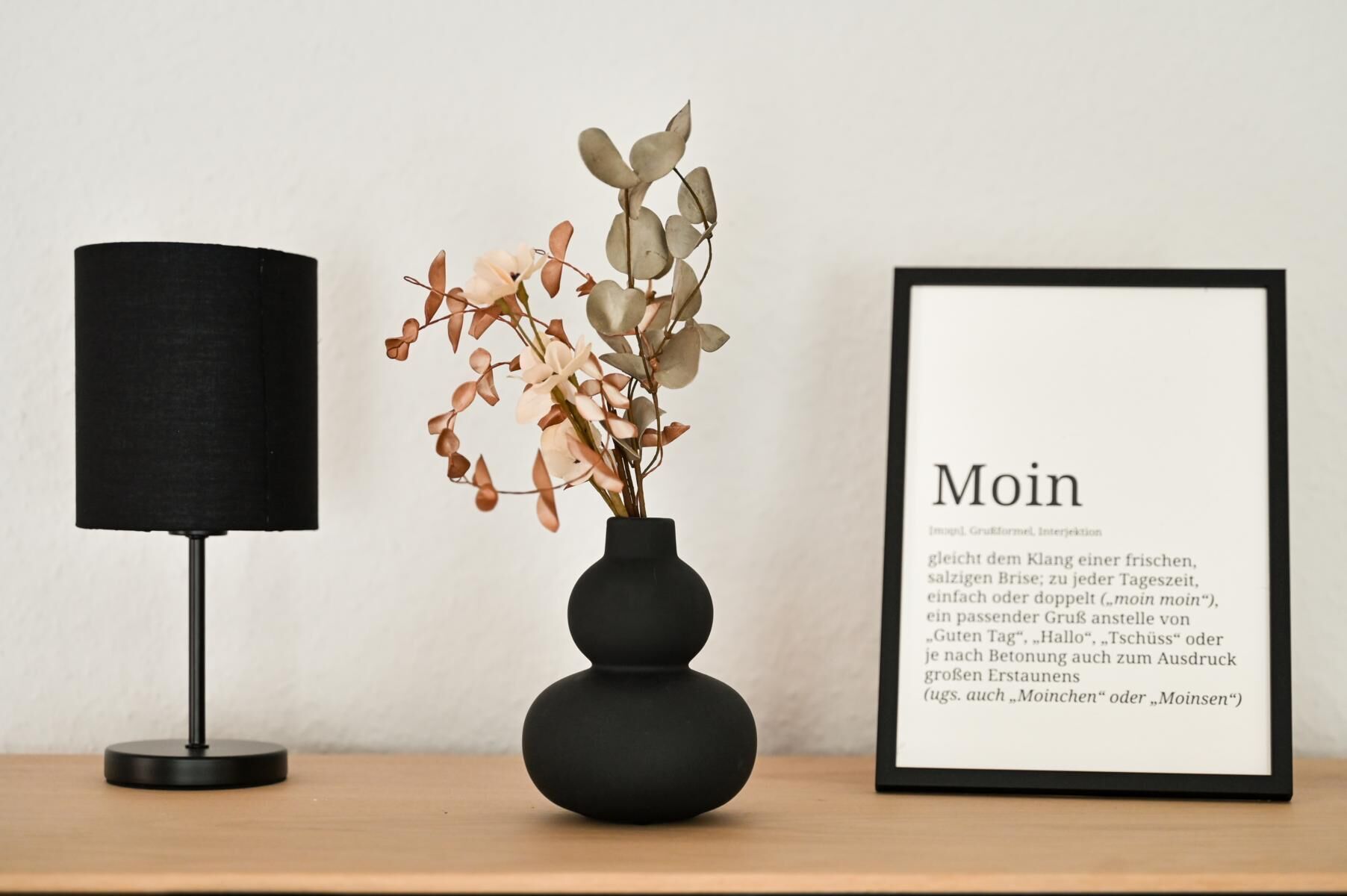 Ein gerahmter Text mit der Aufschrift 'Moin' auf einer Holztischplatte, eine schwarze Lampe und eine Vase mit Blumen.