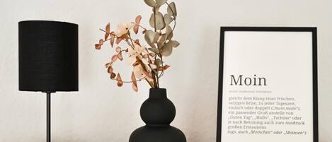Ein gerahmter Text mit der Aufschrift 'Moin' auf einer Holztischplatte, eine schwarze Lampe und eine Vase mit Blumen.