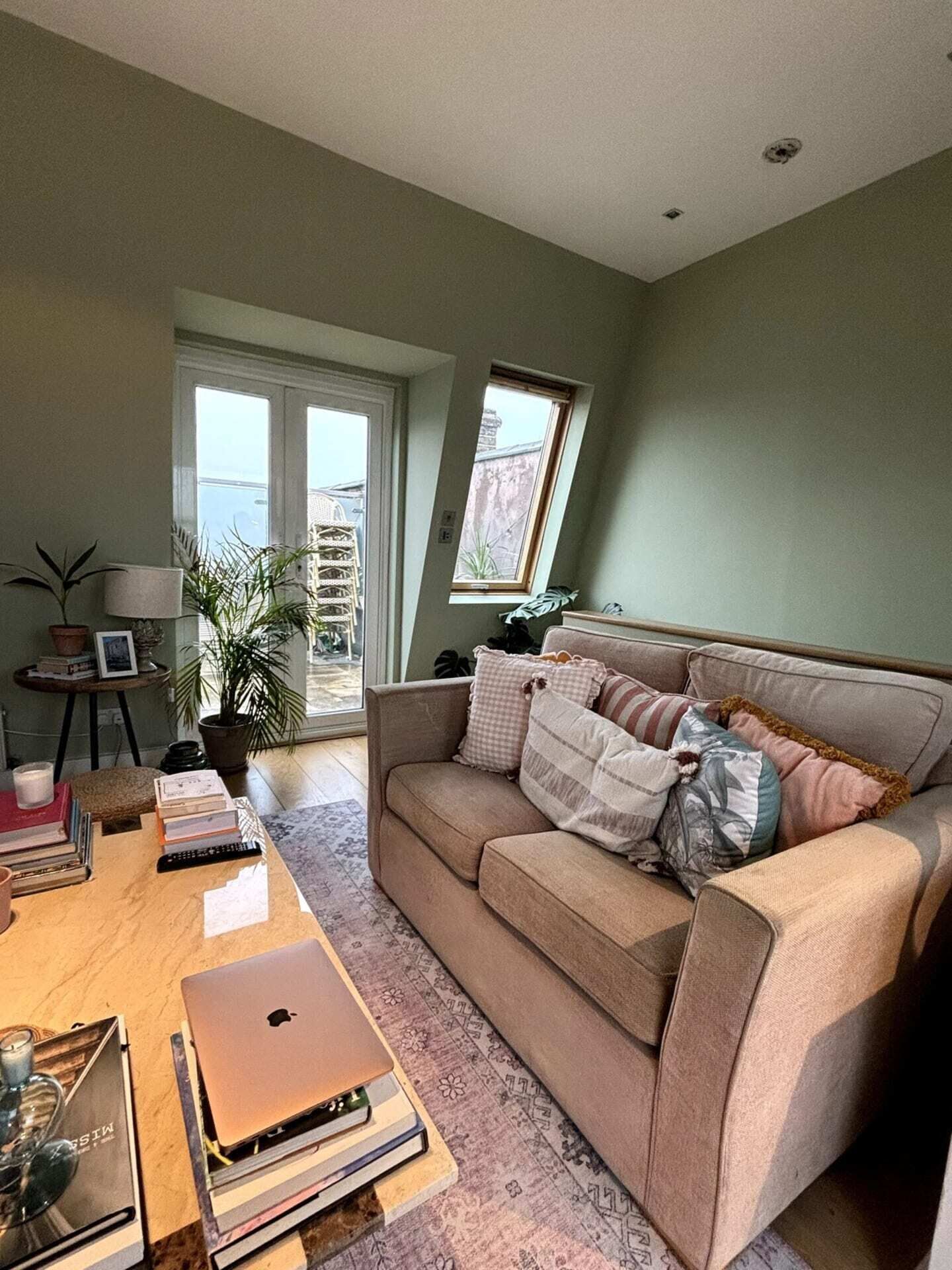 A living room with a beige sofa, a coffee table with books and a laptop, a potted plant, and a view through a glass door.