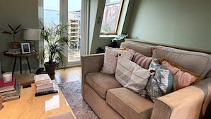 Een woonkamer met een beige bank, een bijzettafeltje met boeken en een laptop, een potplant en uitzicht door een glazen deur.
