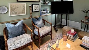 Een woonkamer met twee rieten stoelen, een bijzettafeltje met boeken en een kaars, een boekenkast met diverse spullen, en een piano.