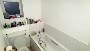 A small, white bathroom with a toilet, a bathtub, and a sink. There is a window above the toilet and a shelf with toiletries. - Cosy Studio Flat - 12 Mins to British Museum! (London)