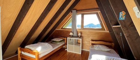 A cozy attic bedroom with two wooden beds, a small desk, and a window overlooking a mountainous landscape.