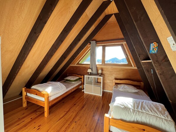 A cozy attic bedroom with two wooden beds, a small desk, and a window overlooking a mountainous landscape.
