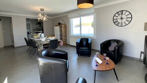 A spacious living room with a dining area, a black leather sofa, a wooden coffee table with wine glasses, and a large wall clock.