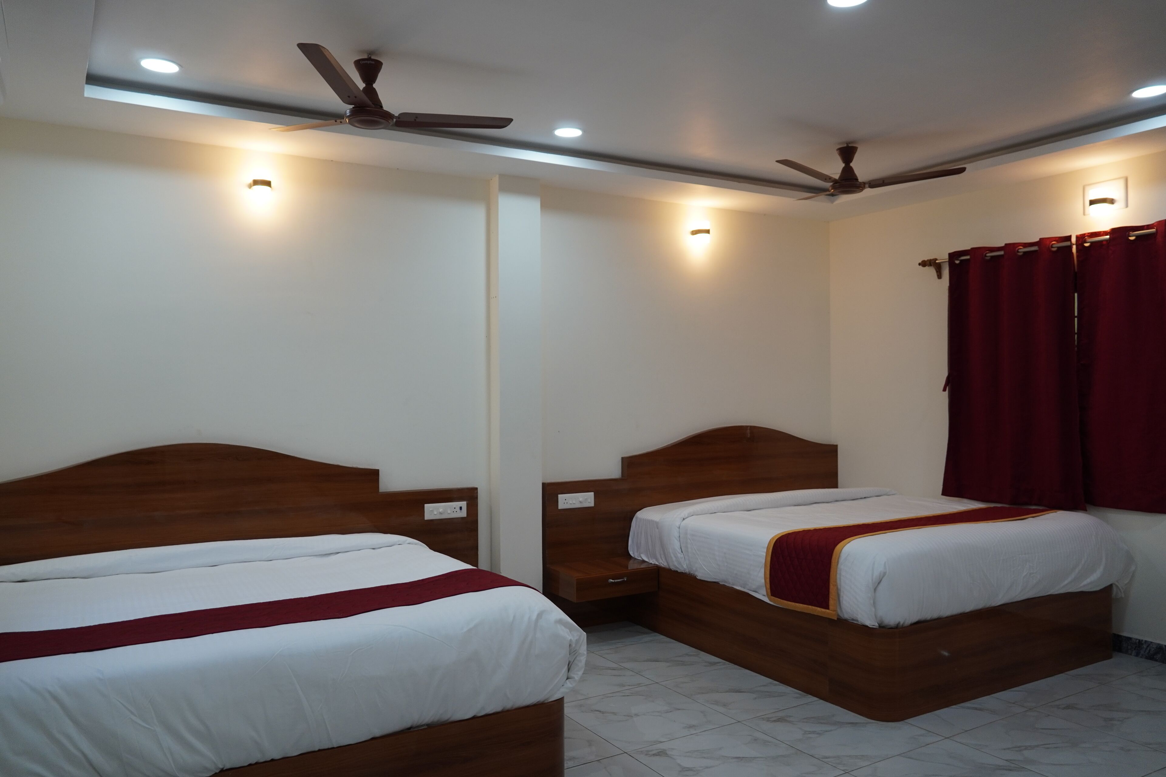 Een hotelkamer met twee houten bedden, een plafondventilator, inbouwverlichting en een rood gordijn.