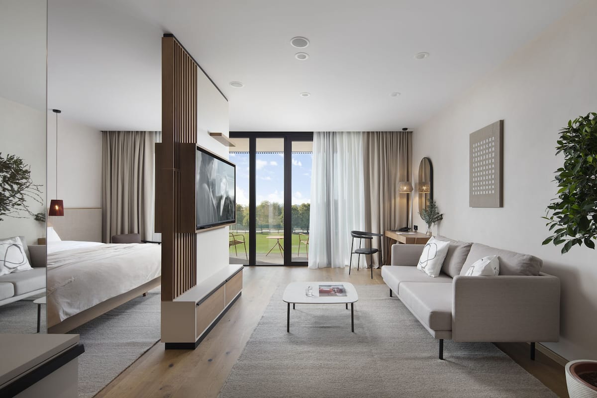 A modern hotel room with a bed, a sofa, a coffee table, and a TV. There is a large window with curtains and a view of the outdoors.