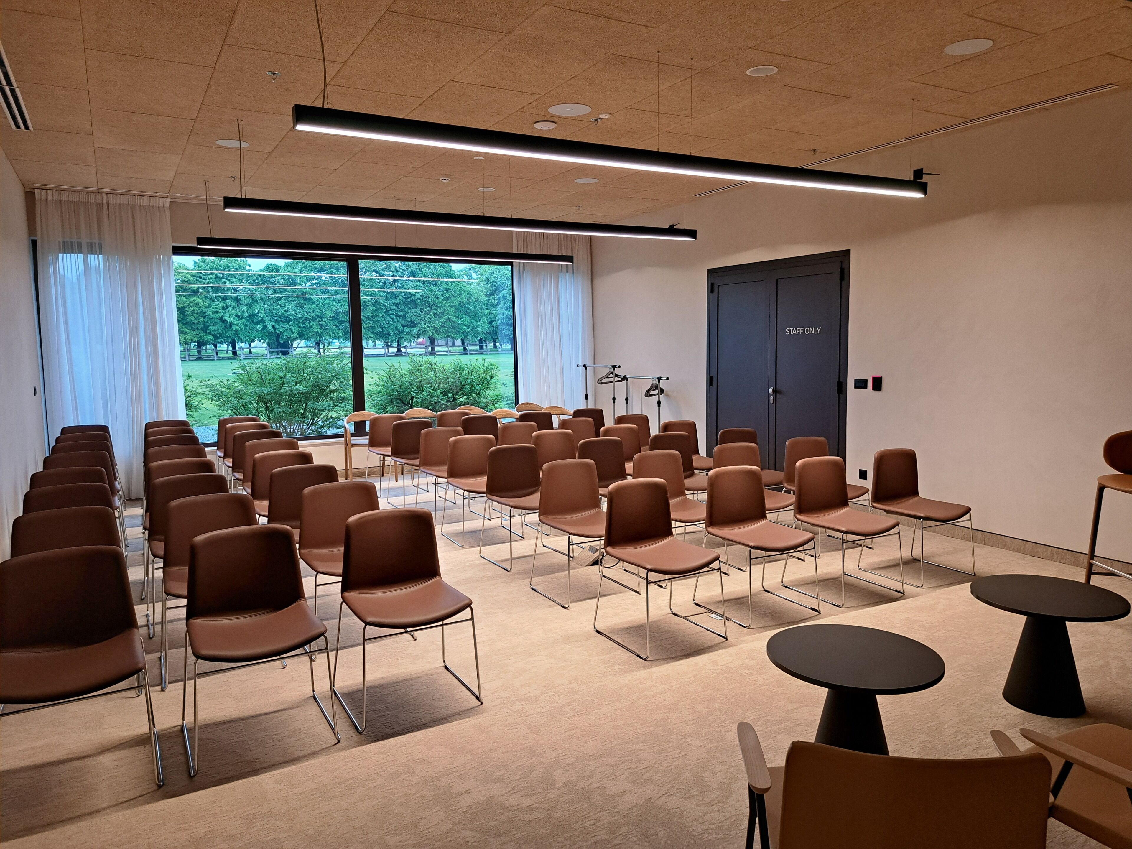 A conference room with rows of chairs and tables, large windows with a view of greenery, and a 'STAFF ONLY' door.