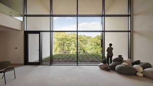 A spacious room with a large window, a person standing by the window, and a seating area with a chair. - Hotel Materra (Cepin)