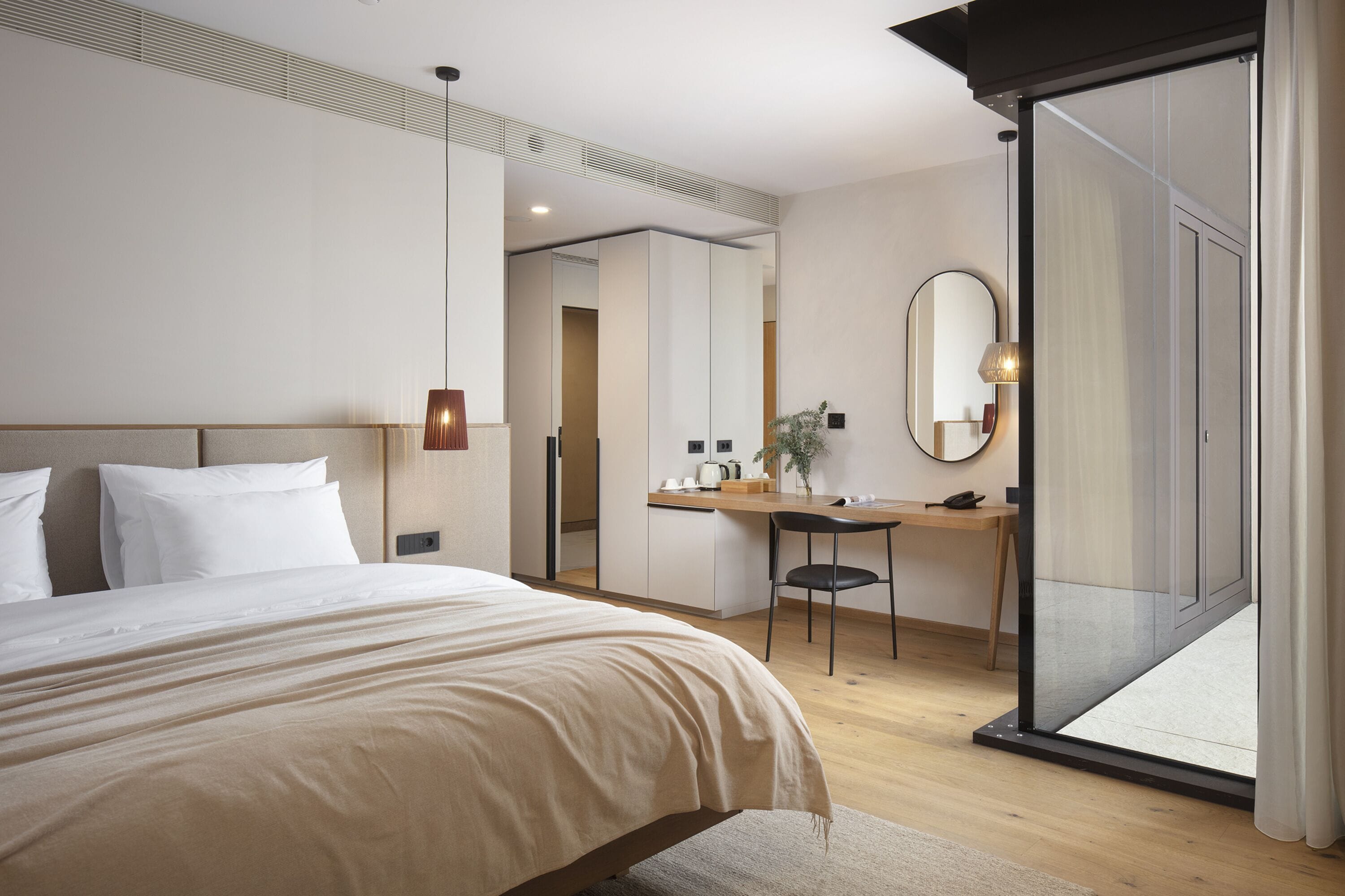 A modern hotel room with a large bed, a desk, a chair, a mirror, and a sliding glass door to the outside.
