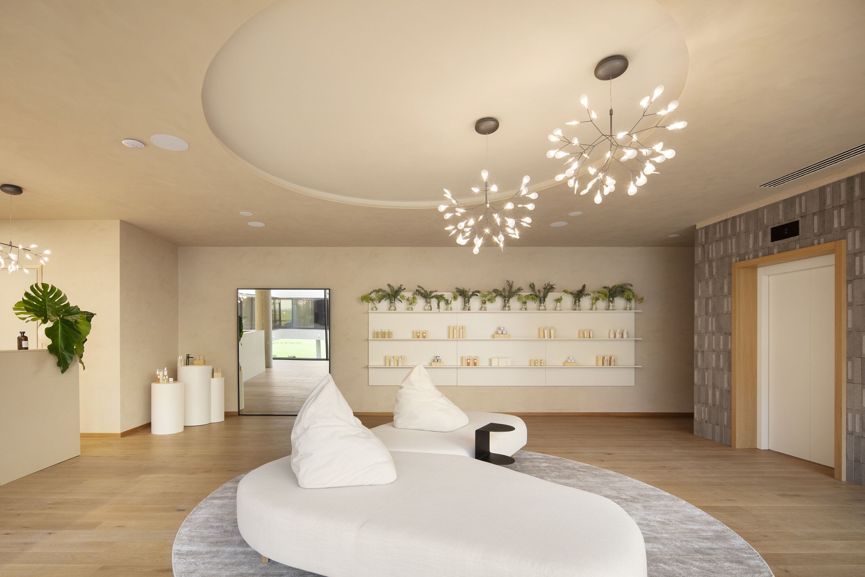A modern interior with a curved white sofa, a ceiling light fixture, and a wall-mounted shelf with various items.