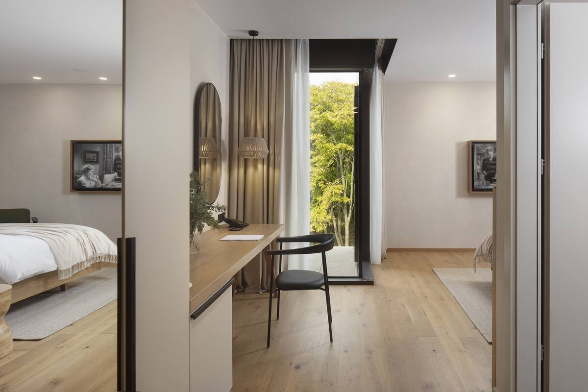 A modern bedroom with a wooden desk, a chair, a bed, and a large window with a view of trees.