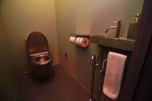 Un baño con inodoro, dispensador de toallas de papel, lavamanos con espejo y una toalla colgada en la pared.