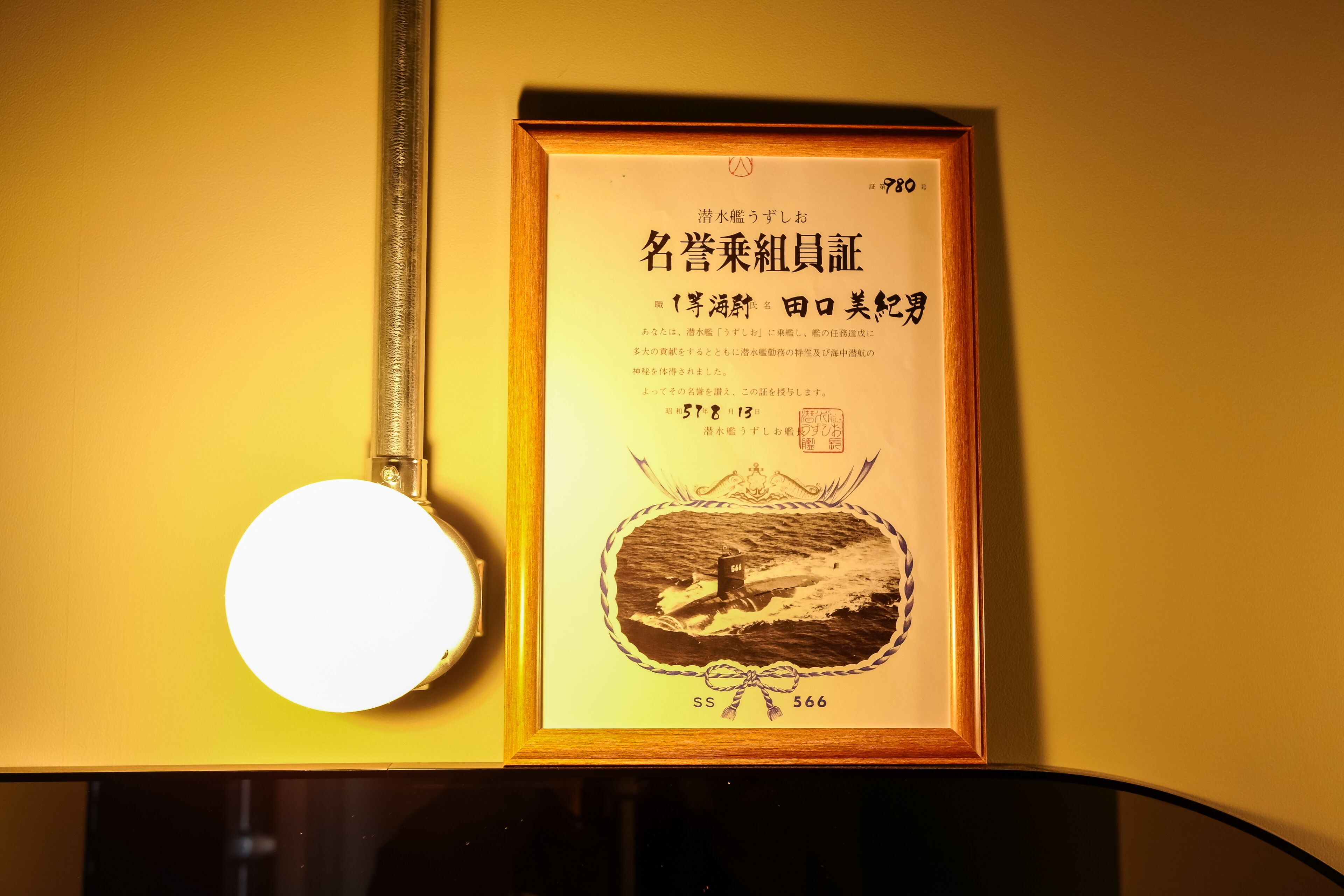 Un certificado enmarco con texto en japonés e ilustración de un barco, colgado en una pared junto a un espejo redondo.