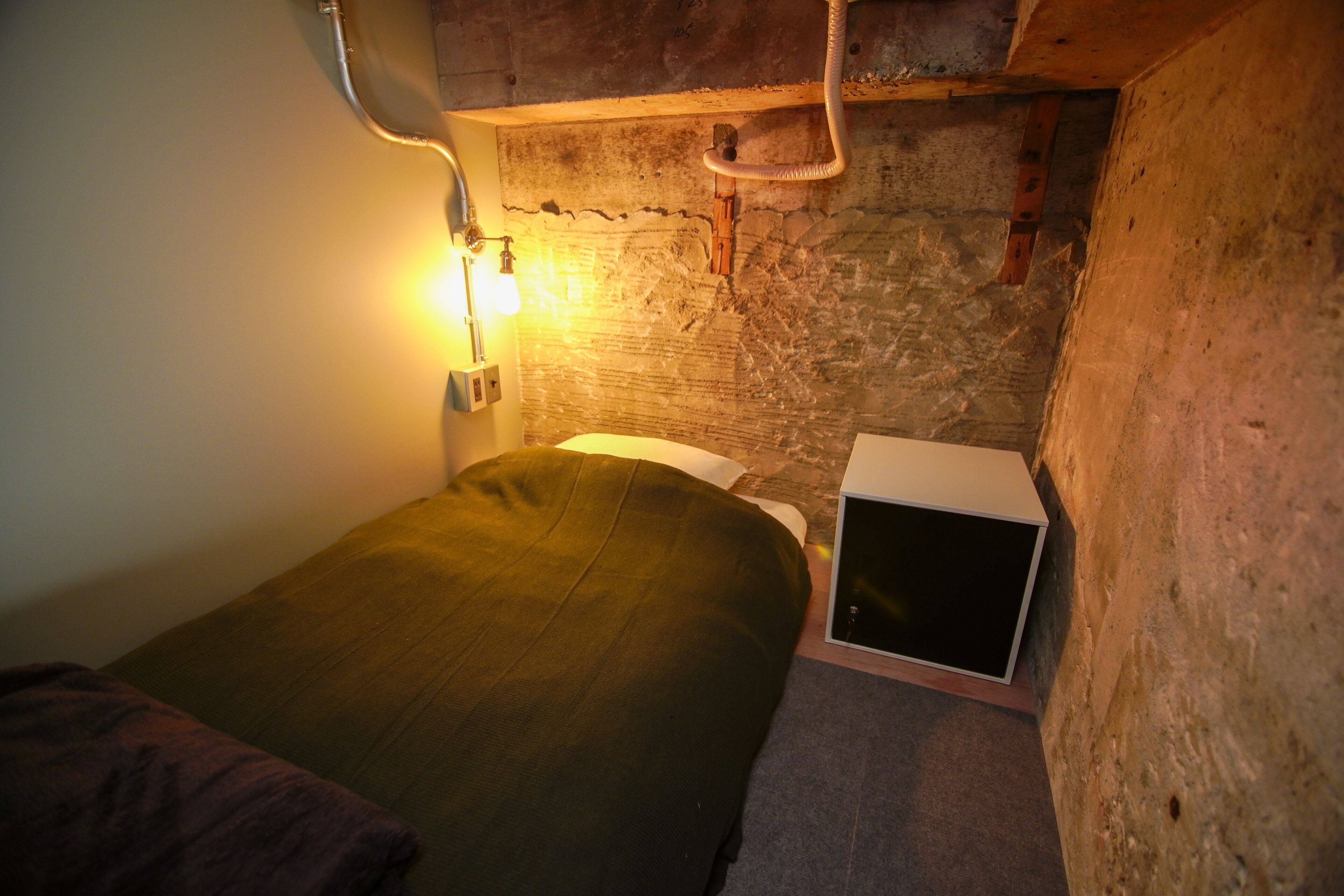 Um quarto pequeno e com pouca luz, com uma cama de solteiro, uma mesa de cabeceira e uma caixa no chão.