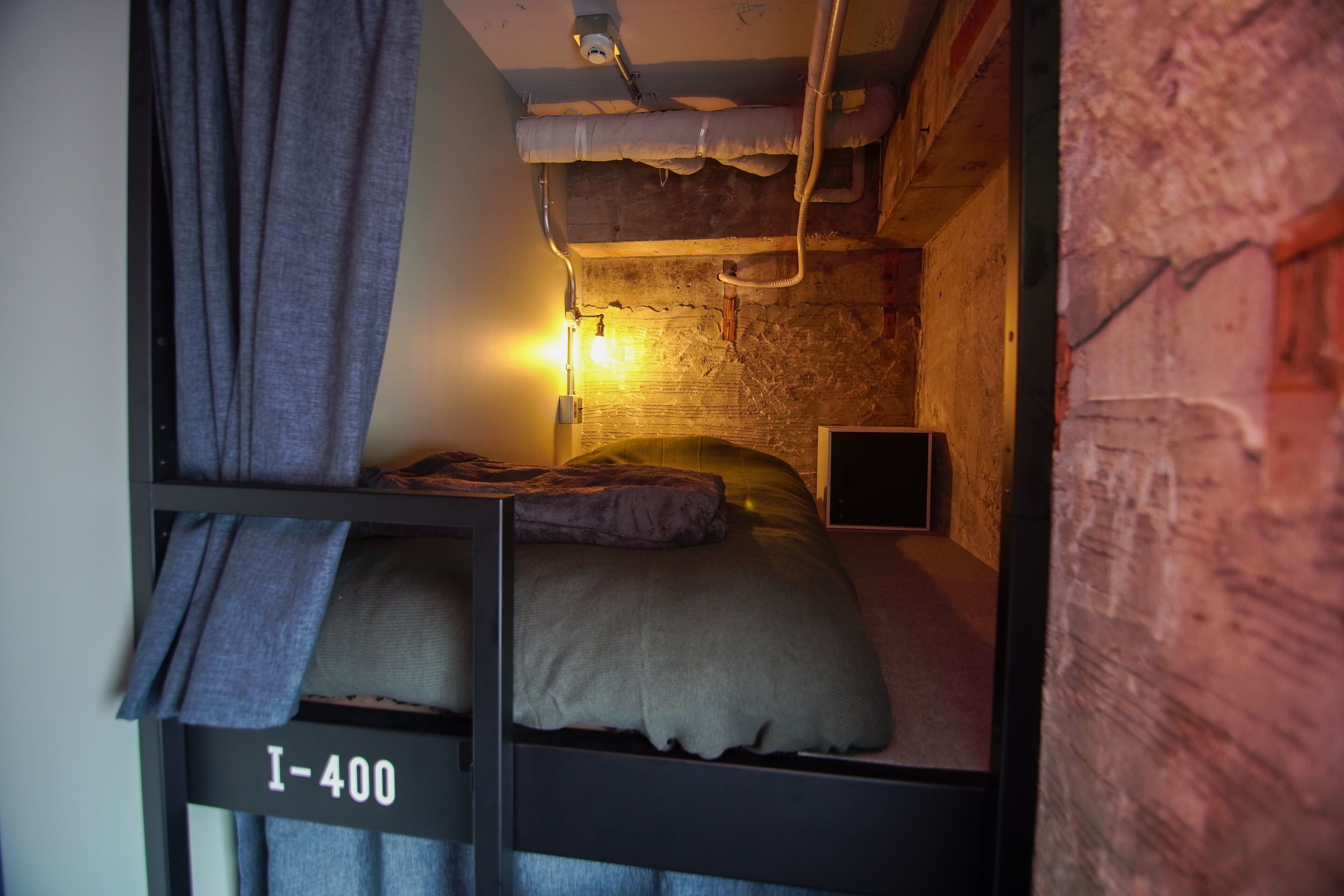 Habitación compacta con una cama litera identificada con la etiqueta “I-400”, una lámpara de pared y paredes de ladrillo visto.