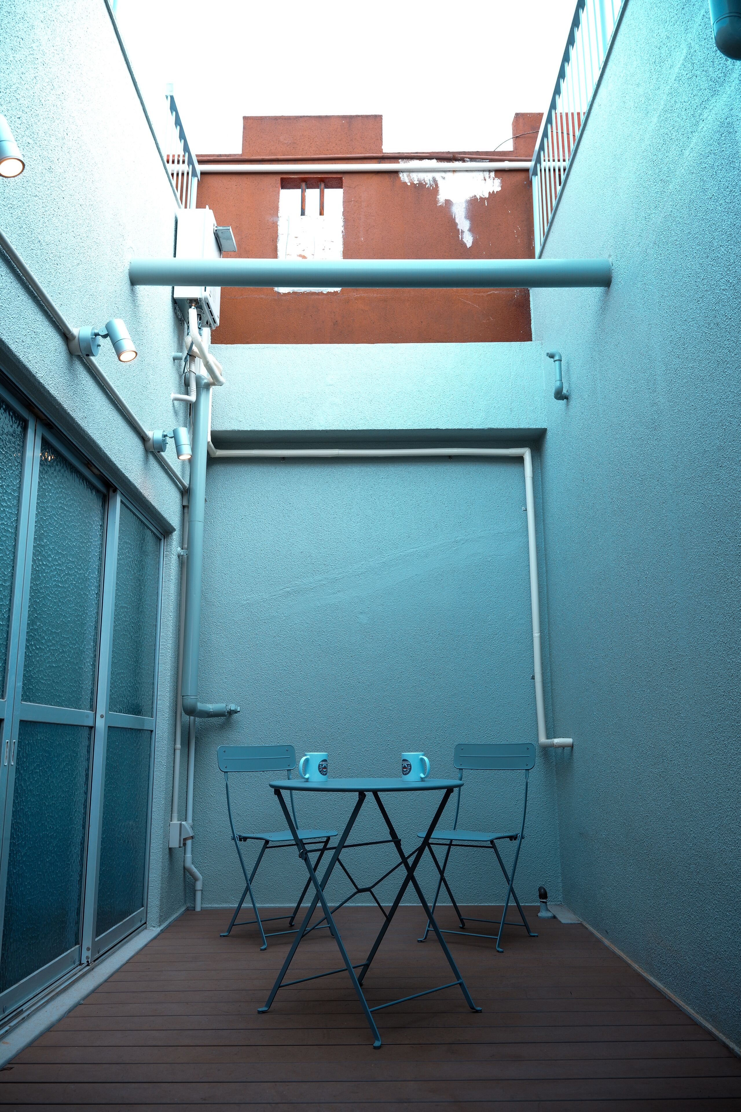 Une petite terrasse extérieure avec deux tables et chaises pliantes en métal, une terrasse en bois et un mur bleu avec une fenêtre et une porte.
