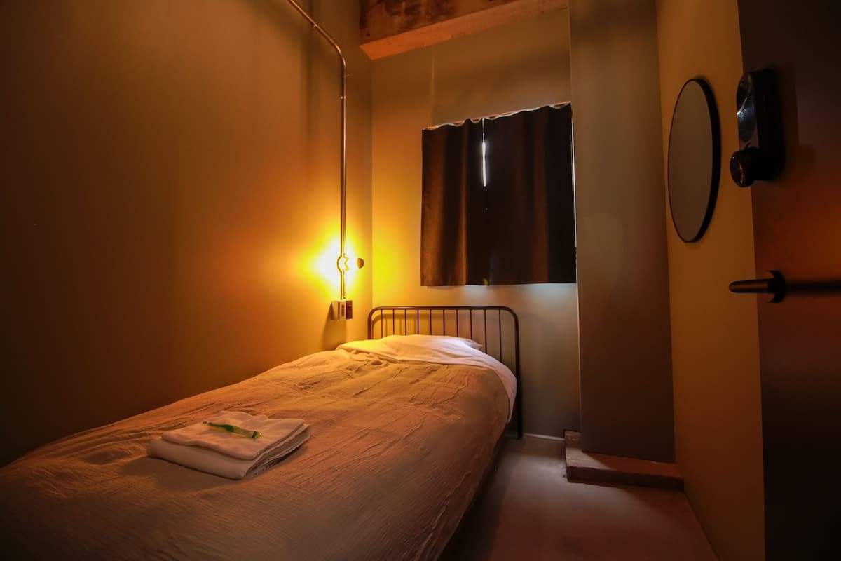 A single bed with a neatly folded white towel and a green toothbrush, a wall-mounted light, a window with a curtain, and a round mirror on the wall.