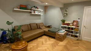 Ein Wohnzimmer mit Fischgrätparkett, einem braunen Sofa, einem kleinen runden Hocker, einem Bücherregal und einer Topfpflanze.