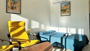 A room with a yellow chair, a wooden table, a blue bed, and two framed pictures on the walls. - Cozy 1BR Apt with Terrace, 150m from Beach, Parking Included - Bormes-les-Mimosas (Bormes-les-Mimosas)