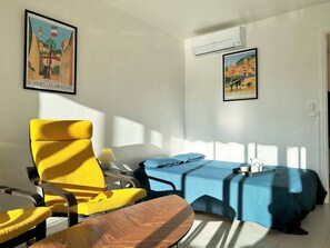Um quarto com uma cadeira amarela, uma mesa de madeira, uma cama azul e duas molduras com quadros nas paredes.
