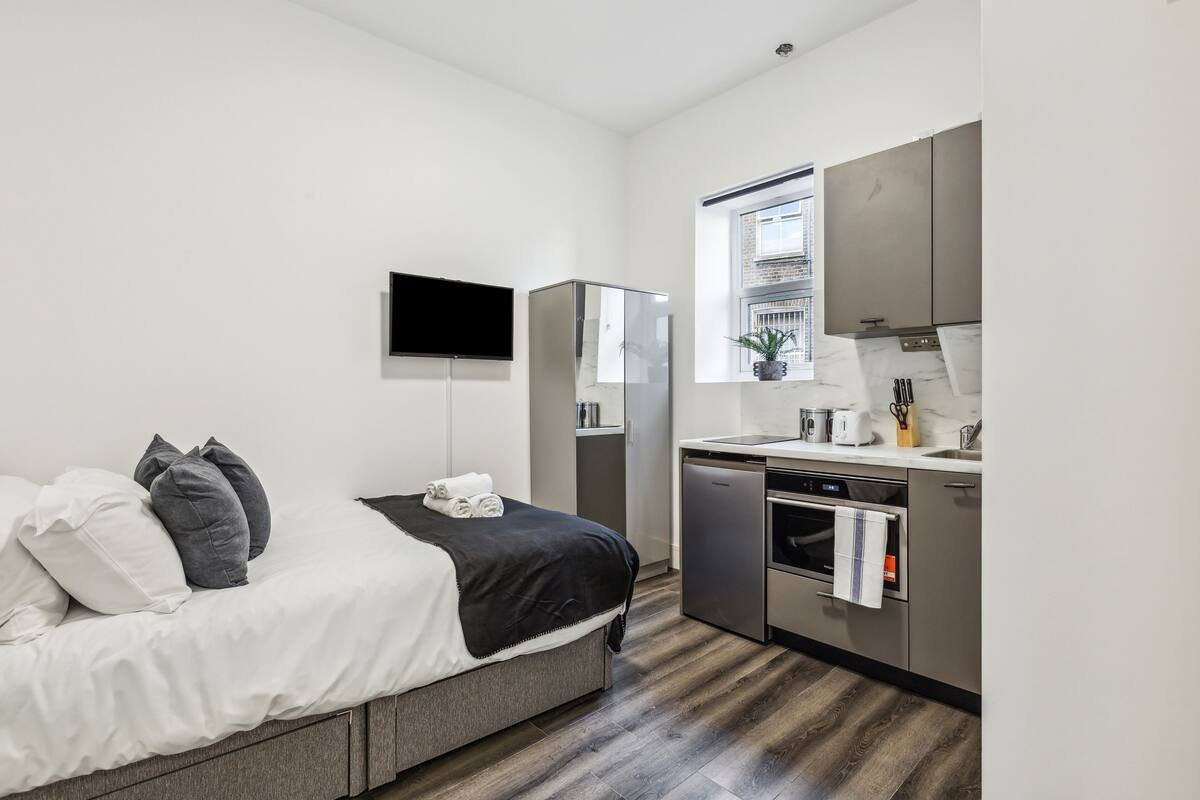 Quarto moderno com cama, televisão de tela plana, guarda-roupa e cozinha equipada com pia e forno.