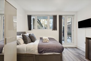 A modern bedroom with a large bed, a TV, and a balcony with a view of the city. - Sutherland Apartments (London)