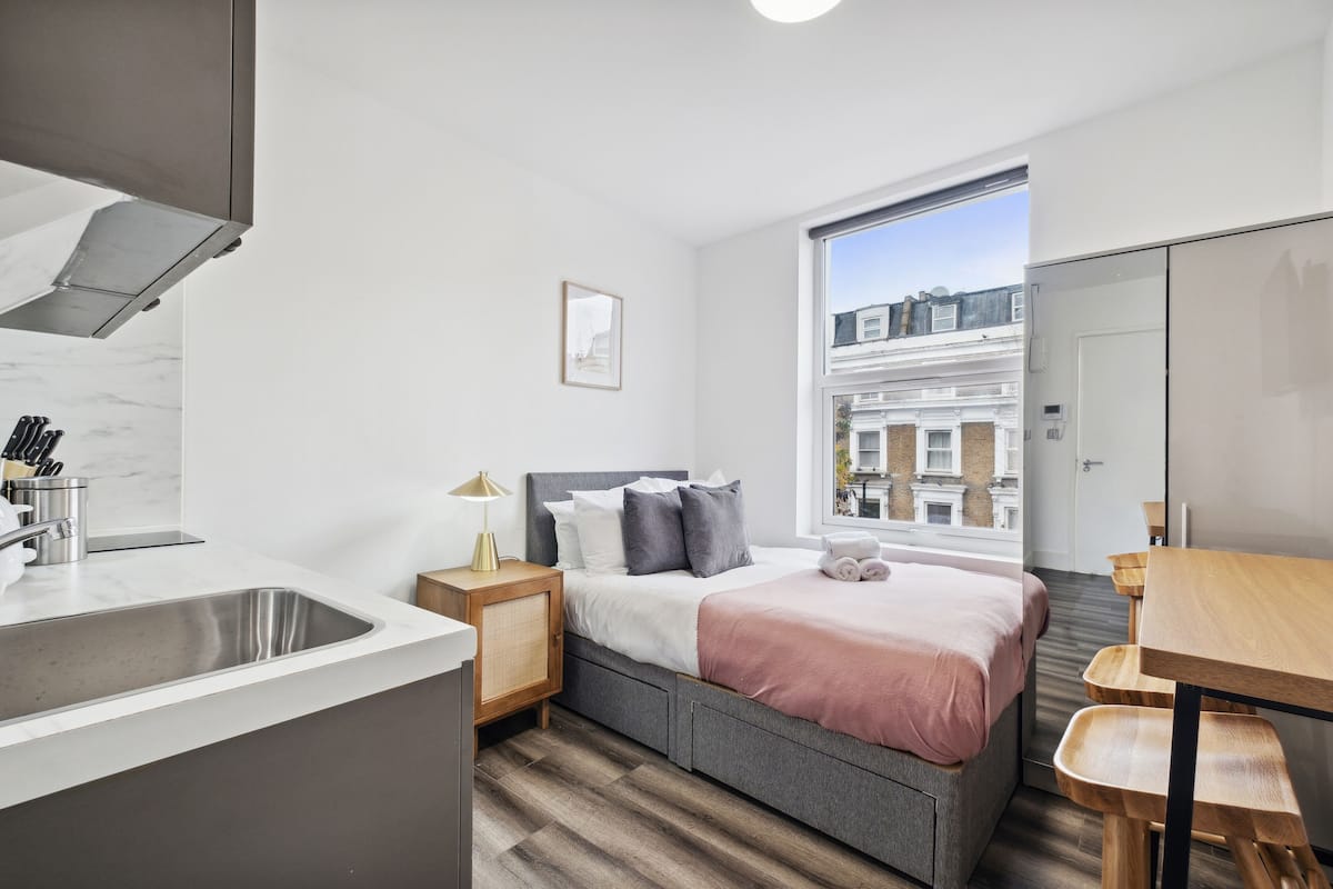 Quarto moderno com cama, mesa de cabeceira e vista para edifícios através de uma janela.