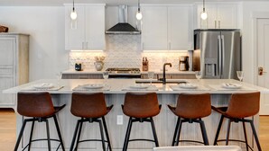 Une cuisine moderne dotée d’un îlot central, de tabourets de bar, d’appareils électroménagers en acier inoxydable, de luminaires suspendus et d’un pare-feu en carreaux.