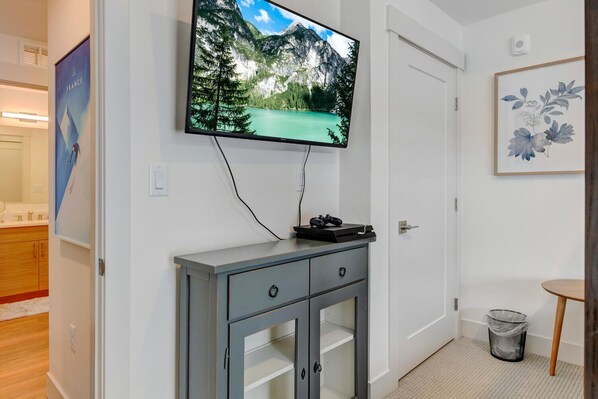 Una habitación con un televisor montado que muestra un paisaje de montaña, un armario gris con cajones, un cesto de basura pequeño y una obra de arte floral enmarcada en la pared.