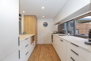 A modern kitchen with white cabinetry, a wooden accent wall, a built-in oven, and a window overlooking a brick wall. - Inn Sophia - Two Homes, Spa Pools, Sleeps 10 (Rotorua)