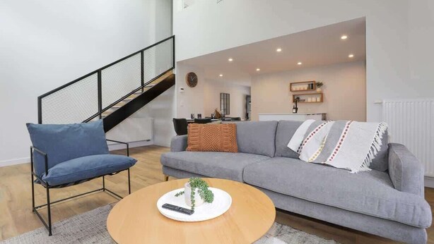 Una sala moderna con un sofá gris, un sillón azul y una mesa de centro de madera. Se ve una escalera en el fondo que conduce a un nivel superior.