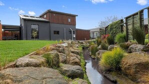 A modern building with a flat roof and large windows, surrounded by a landscaped garden with a small water feature. - Inn Sophia - Two Homes, Spa Pools, Sleeps 10 (Rotorua)