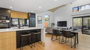A modern kitchen with a central island, bar stools, and a dining area with a large window. - Inn Sophia - Two Homes, Spa Pools, Sleeps 10 (Rotorua)