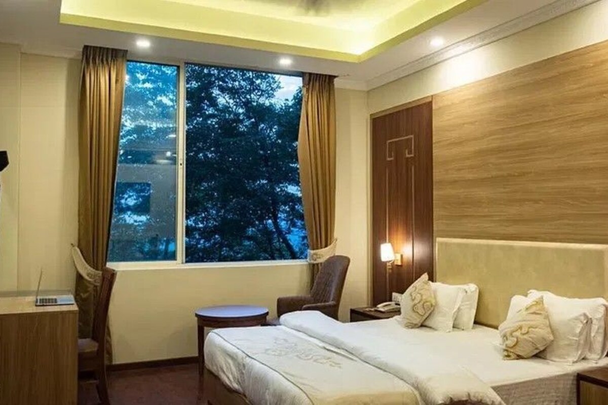 A hotel room with a bed, a desk, a chair, a window with curtains, and a nightstand with a lamp.