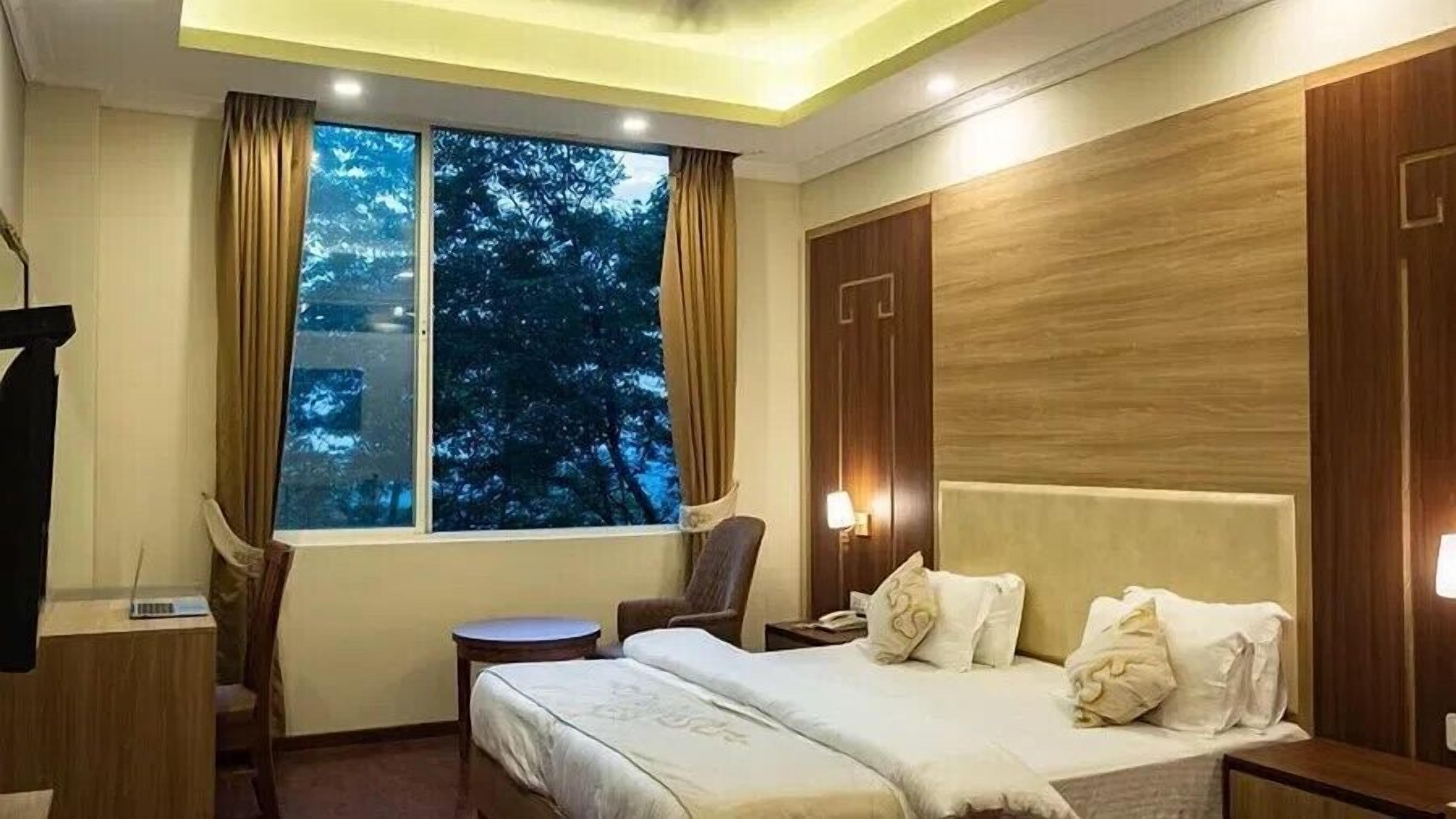 A hotel room with a bed, a desk, a chair, a window with curtains, and a nightstand with a lamp.