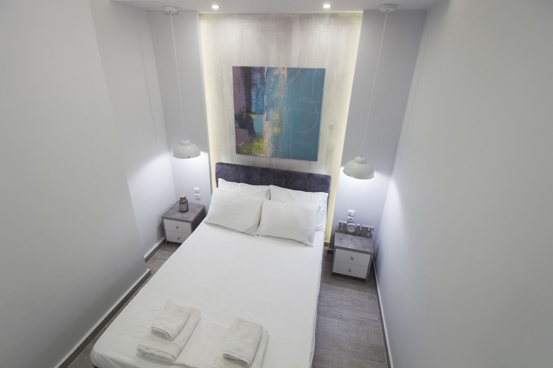 A hotel room with a large bed, two bedside tables, a painting above the bed, and two wall-mounted lamps.
