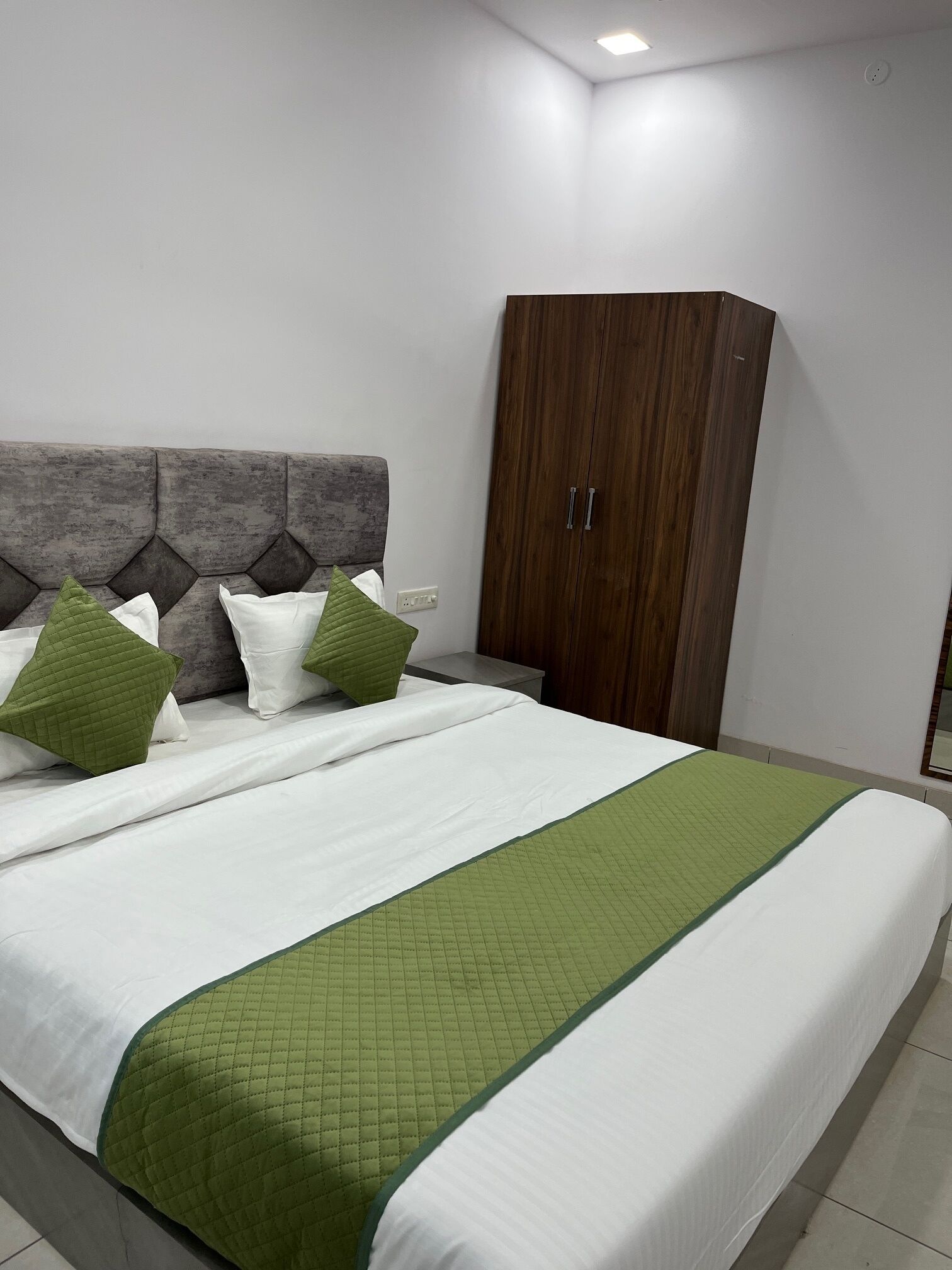 A neatly made bed with a green quilted bedspread and white linens, a grey upholstered headboard, and a wooden wardrobe with two doors.
