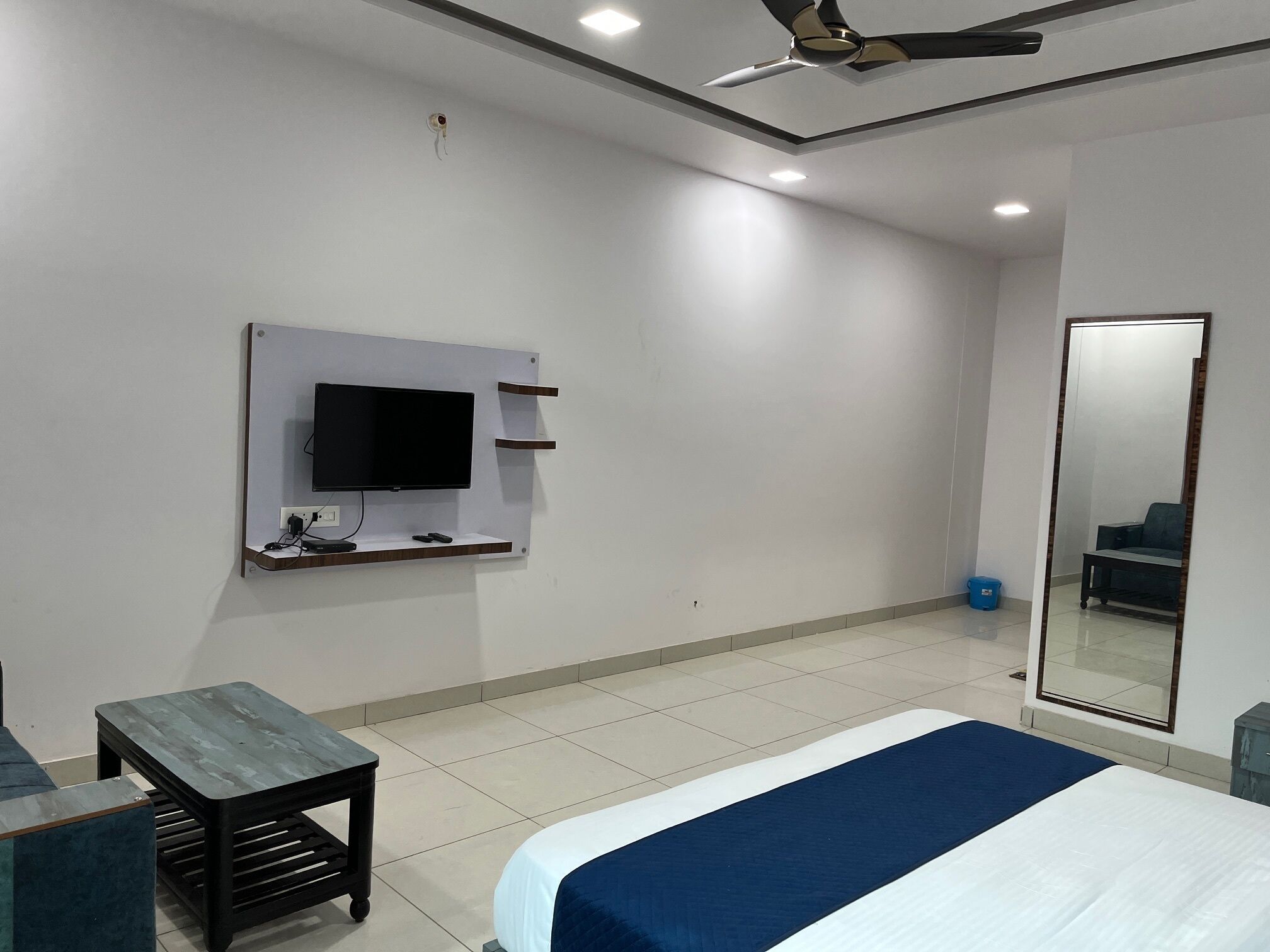 Una habitación de hotel moderna con una cama grande, un televisor de pantalla plana montado en la pared, una mesita pequeña y un ventilador de techo.