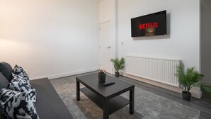 A modern living room with a flat-screen TV displaying "NETFLIX" in red letters, a black coffee table with a remote control, a grey sofa, and two potted plants. - Isimi Luxurious 4 bed 4 en -suite House Cumbria (Barrow-in-Furness)