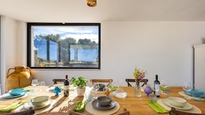 A dining area with a large window, wooden table, chairs, and various table settings including plates, glasses, and a vase with flowers. - 3670 Villa Perlaianca by Perle di Puglia (Carovigno)