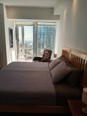 Une chambre d’hôtel moderne avec un grand lit, un fauteuil et une vue sur le paysage urbain, visible à travers une grande fenêtre.