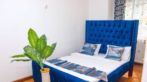 A bedroom with a blue upholstered headboard, a bed with white bedding and patterned pillows, a large green plant in a striped vase, and a wooden floor. - Lux Suites Embakasi Airport Apartments (Nairobi)