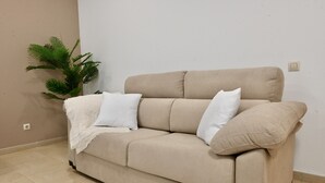 A beige sofa with white cushions and a white throw blanket, positioned against a plain wall with a potted plant to the left.